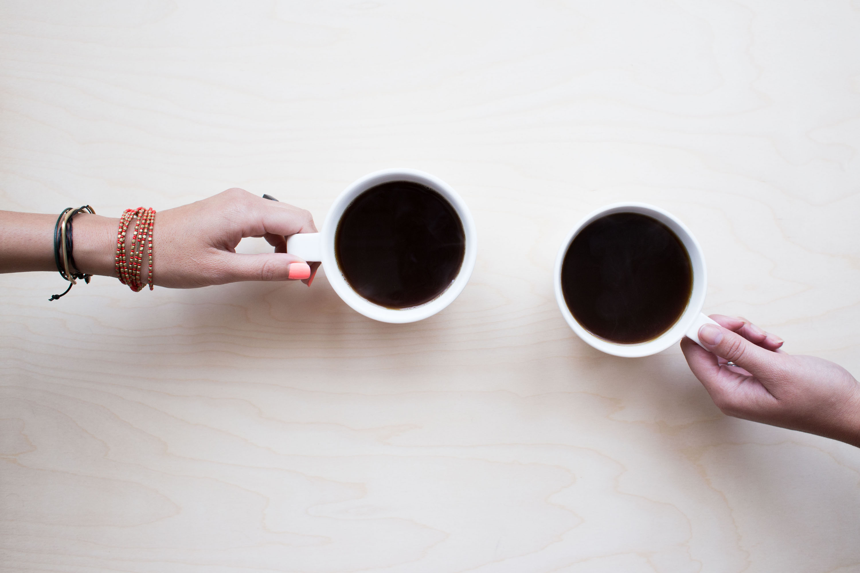 Two hands hold two coffee cups on a wooden table.